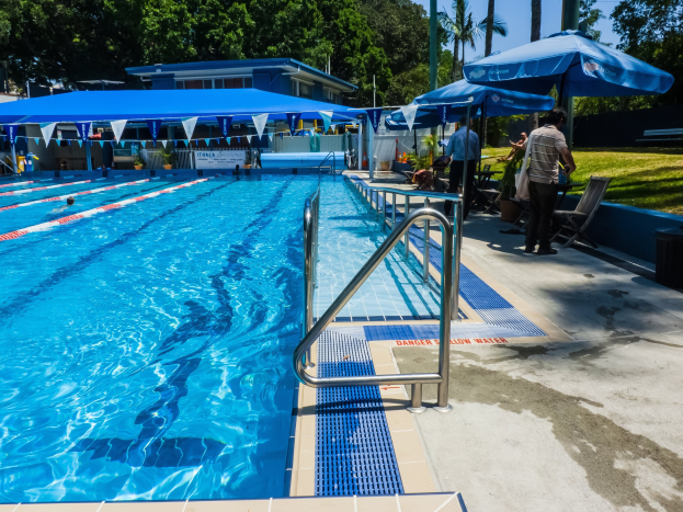 Großer rechteckiger Swimmingpool mit schwimmenden Menschen, umgeben von Geländern, Sonnenschirmen, Stühlen, Tischen, Pflanzen und Bäumen, mit einem Gebäude im Hintergrund unter einem klaren blauen Himmel.