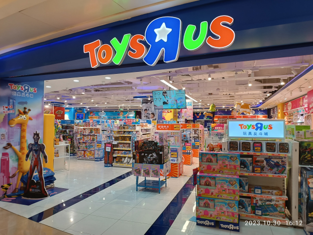 Ein Toys R Us-Laden in einem Einkaufszentrum mit Regalen voller Spielzeug, einer Schildertafel mit Text, Deckenleuchten und einem Wasserzeichen in der rechten unteren Ecke.