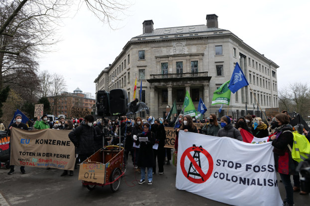 Große Gruppe von Menschen bei einer Straßenprotest gegen fossile Brennstoffe, die Schilder und Fahnen tragen, mit einem Fahrzeug im Vordergrund und Gebäuden, Bäumen und einem klaren blauen Himmel im Hintergrund.