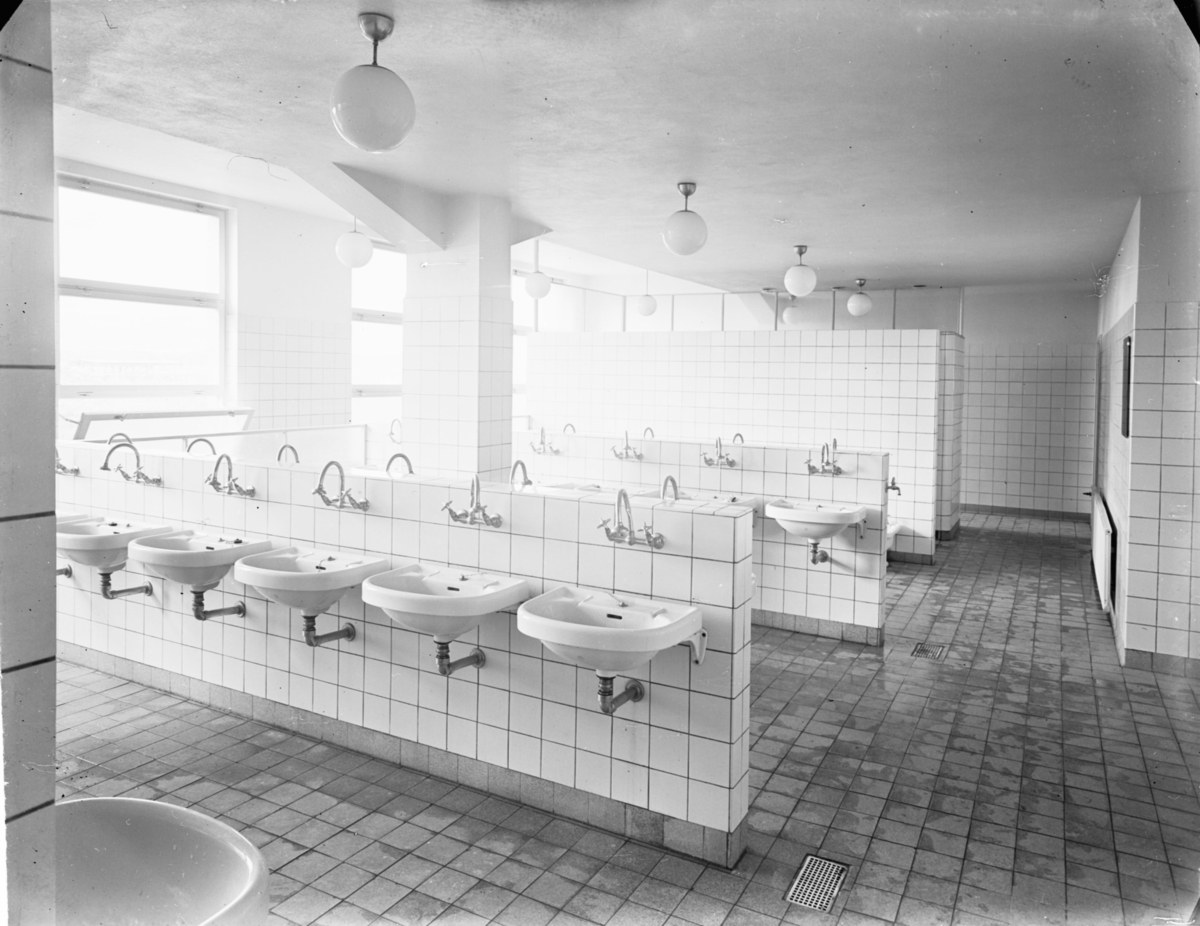 Schwarzes und weißes Foto einer öffentlichen Toilette mit Waschbecken, Wasserhähnen, Wänden, Beleuchtung und Fenstern.