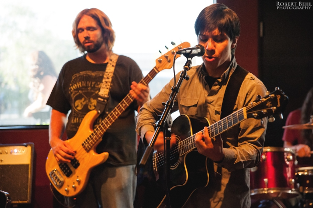 Eine Person, die auf einem Boden steht und eine Gitarre hält und singt, mit einer anderen Person, die Gitarre spielt, und Trommeln im Hintergrund.