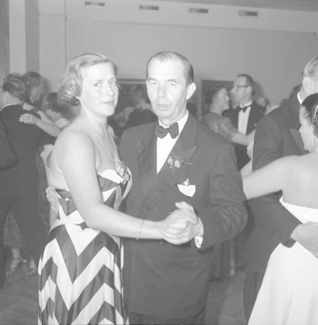 Ein Mann in einem schwarzen Blazer, weißem Hemd und schwarzen Hosen tanzt mit einer Frau in unbekannter Kleidung auf einer formellen Veranstaltung, indem sie sich an den Händen halten und zur Musik wiegen.