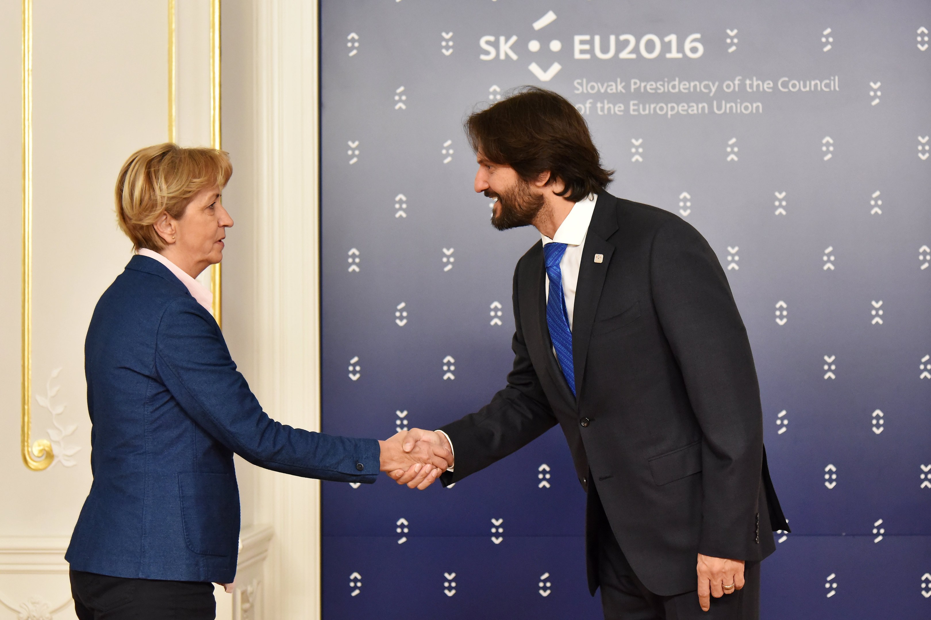 Bundeskanzlerin Angela Merkel und Bundespräsident Joachim Gauck schütteln Hände vor einer Wand mit einem "EU-Gipfel"-Banner und einer Tür im Hintergrund, beide lächeln.