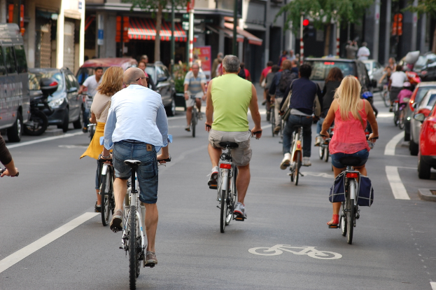 Menschen fahren Fahrrad auf Straßen mit Autos auf der linken und rechten Seite, sowie Geschäfte, ein Zelt, Bäume und Gebäude im Hintergrund.