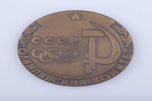 Eine Bronzemedaille mit der Aufschrift "Olympische Spiele" und einem Hammer und Sichel-Symbol auf einem weißen Hintergrund.