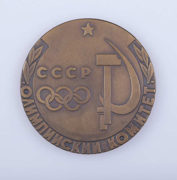 Eine Bronzemedaille mit der Aufschrift "Olympische Spiele" und einem Hammer und Sichel-Symbol auf einem weißen Hintergrund.
