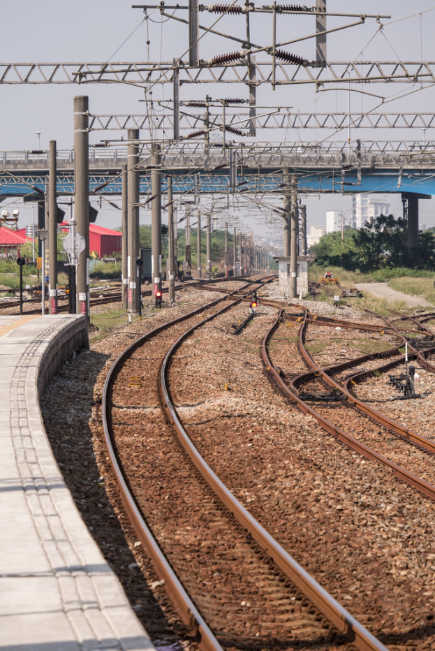 Eine Plattform auf der linken Seite, Eisenbahnschienen auf der rechten Seite, mit Pfählen, einer Brücke, Bäumen, Gebäuden und bewölktem Himmel im Hintergrund.