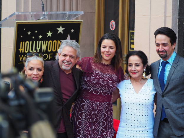 Eine Gruppe von Menschen steht vor einem Hollywood Walk of Fame-Star, mit einem Podium und einem Mikrofon in der Mitte, einer Tafel mit Text dahinter und einer Person mit einer Kamera links, mit Wänden und einer Tür im Hintergrund.