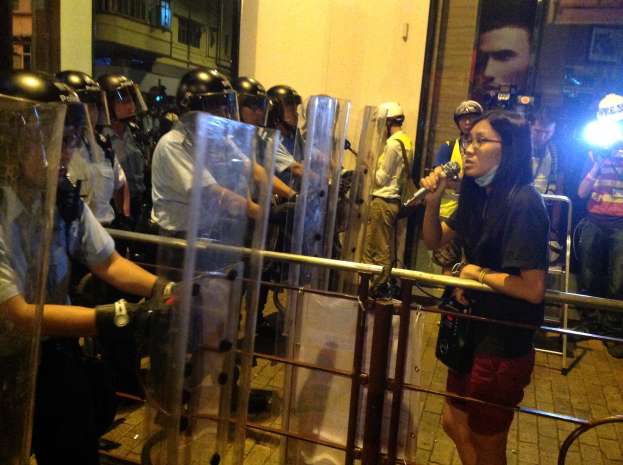 Eine Frau in einem schwarzen T-Shirt und roten Shorts steht vor der Bereitschaftspolizei, ein Mikrofon haltend, mit einer Menge, einer Barriere und Gebäuden im Hintergrund.