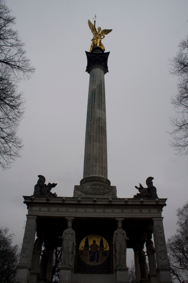 Ein Denkmal mit Statuen und Säulen im Vordergrund, trockene Bäume rechts, grüne Bäume links, unter einem bewölkten Himmel.