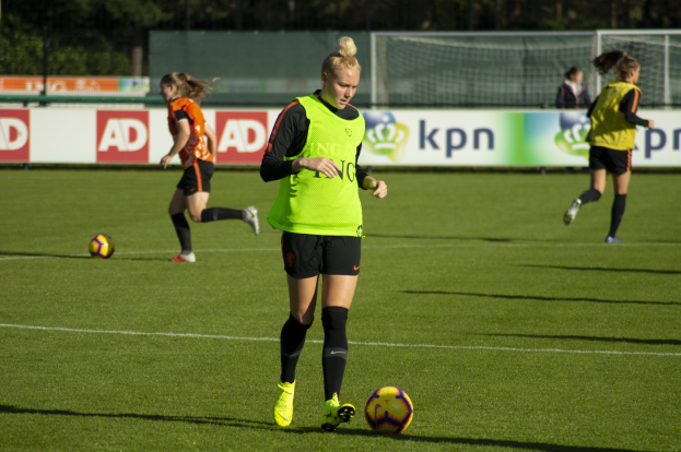 Eine Gruppe von Frauen in Fußballtrikots, die auf einem Feld mit Bäumen im Hintergrund Fußball spielen. Banner mit Text und ein Netz im Vordergrund.
