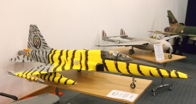 Verschiedene Flugzeugmodelle sind auf Tischen mit Etiketten angeordnet, ein Stuhl befindet sich links und eine Wand im Hintergrund.