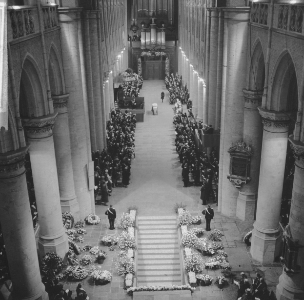 Schwarz-weiß-Foto einer Trauerfeier in einer Kirche mit Menschen auf dem Boden, Blumensträuße auf beiden Seiten der Treppe zum Altar, Säulen, Lichter und einem Kronleuchter.