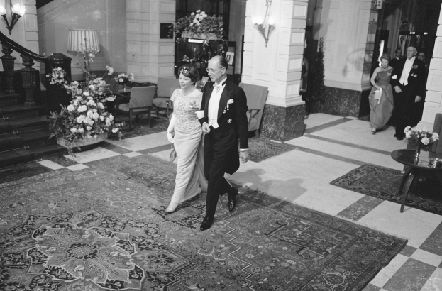 Schwarz-weiß-Foto einer Braut und eines Bräutigams, die durch die Hotellobby gehen und von Gästen umgeben sind, mit Blumenschmuck, Möbeln und architektonischen Details.