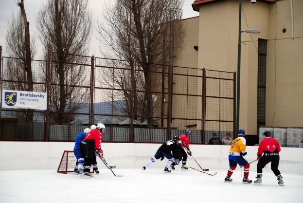 Personen beim Eishockey auf einem Eisstadion mit Gebäuden, Bäumen, einer Straßenlaterne, einem Namensschild und Zäunen im Hintergrund unter einem Himmel.