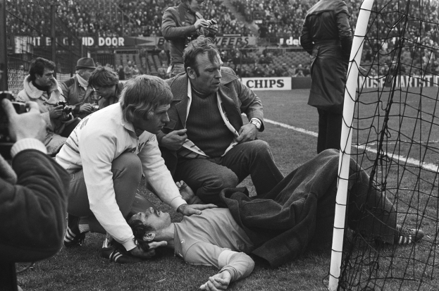 Ein Mann liegt auf dem Boden in der Nähe eines Fußballtors, umgeben von sitzenden und stehenden Zuschauern auf dem Rasen, mit einem Netz auf der rechten Seite und einer Menge in den Tribünen dahinter.