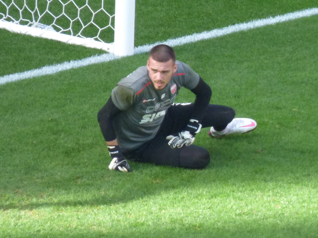 Ein Fussballspieler kniet vor einem Tor, trägt eine Sportkleidung, Handschuhe und Turnschuhe und hat den Torpfosten im Hintergrund sichtbar.