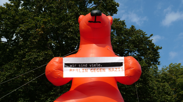 Aufblasbarer Bär mit einem Schild "Berlin gegen Nazis" vor Bäumen und Wolken.