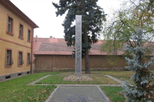 Ein Denkmal steht auf einer grasbewachsenen Fläche neben einem Baum, umgeben von Gebäuden mit Fenstern, einem Pfad und trockenen Blättern auf dem Boden, unter einem bewölkten Himmel und ruft eine traurige Stimmung hervor, die an ein Mahnmal für Auschwitz-Opfer erinnert.