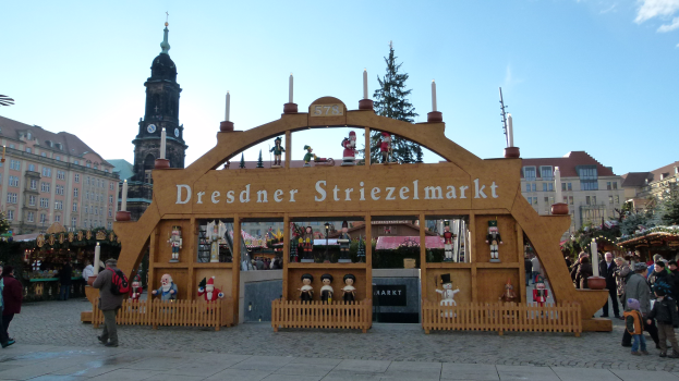 Ein lebendiger Weihnachtsmarkt in Dresden, Deutschland, mit Menschen, die herumlaufen, einige mit Taschen, und einem Bogen mit Text darauf, vor einem Hintergrund aus Bäumen, Gebäuden mit Fenstern und einem bewölkten Himmel.
