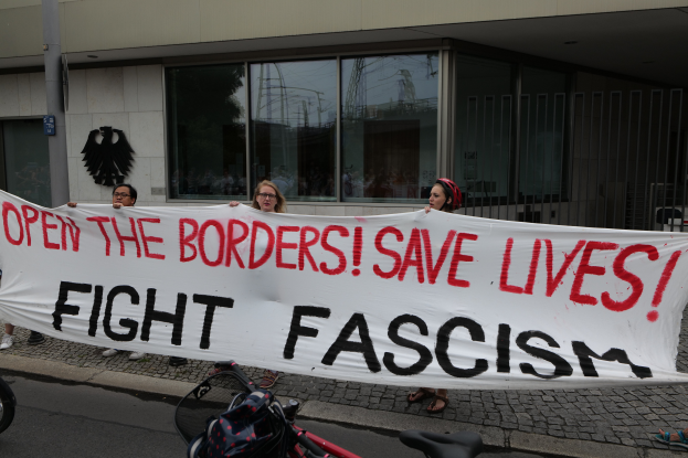 Eine Gruppe von Menschen hält ein Transparent mit der Aufschrift 'Öffnet die Grenzen, rettet Leben, kämpft gegen Faschismus' vor einem Gebäude mit Glasfenstern und einem Mast im Hintergrund, mit einem Fahrrad und einer Tasche auf der Straße.