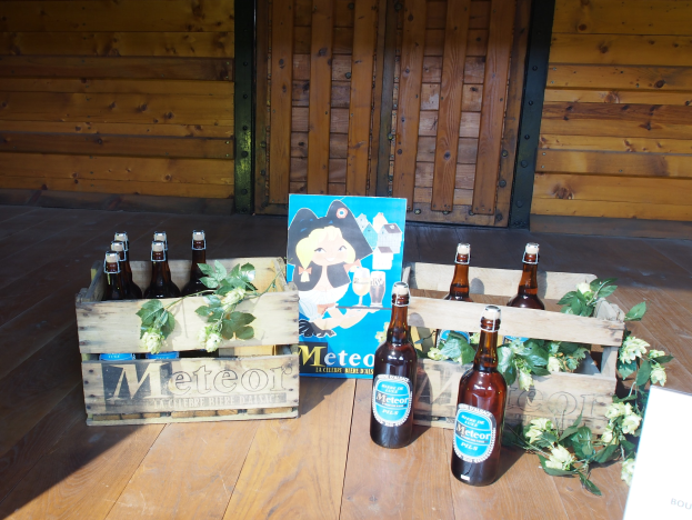 Ein hölzerner Kasten voller Bierflaschen auf einem hölzernen Boden in einer Gartenumgebung, umgeben von Blättern und Blumen, und eine hölzerne Wand und eine Tür im Hintergrund.