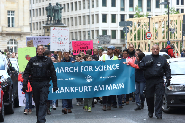 Eine Gruppe von Menschen marschiert mit einem Banner "March for Science Frankfurt am Main" die Straße entlang, während Autos daneben fahren und Gebäude, Statuen, Laternenpfähle, Schilder und Bäume im Hintergrund zu sehen sind.