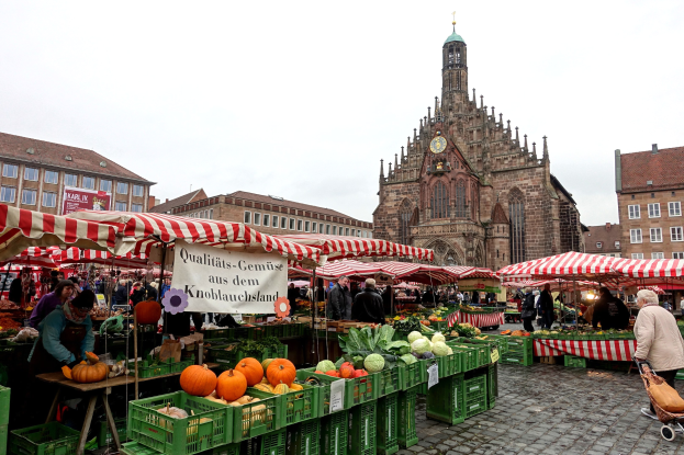 Ein belebter Markt in Nürnberg, Deutschland, mit Obst, Gemüse, Zelten, Menschen, Gebäuden, einem Kirchturm und Himmel.