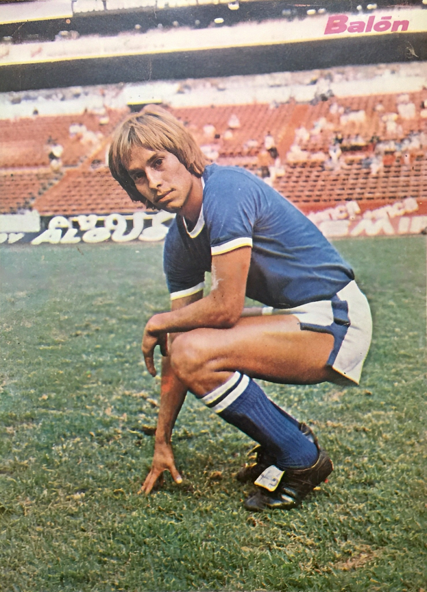 Ein Mann kniend auf einem Fußballfeld umgeben von Menschen mit Schildern, trägt Sportschuhe.