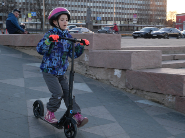 Ein junger Junge in Helm und Handschuhen fährt auf einem Roller eine Treppe hinunter, vorbei an Fahrzeugen, Menschen, Bäumen, Pfosten, Brettern, Gebäuden und einem klaren blauen Himmel.