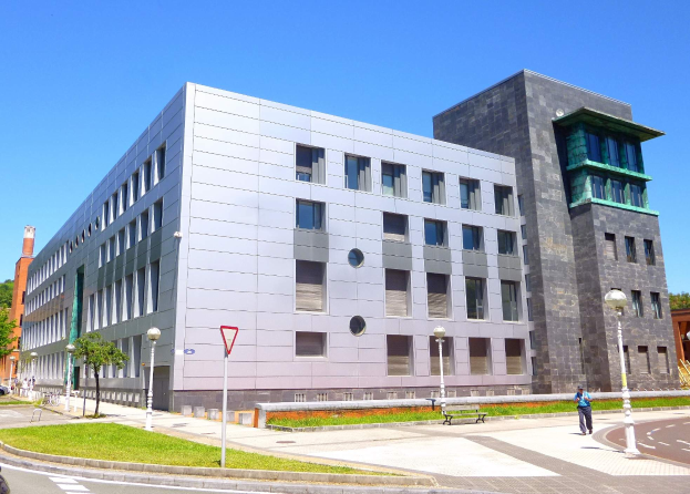 Die Universität Helsinki, ein großes Gebäude mit zahlreichen Fenstern, ist von Straßenlaternen, Autos, Fußgängern, Bäumen und einem klaren blauen Himmel umgeben.