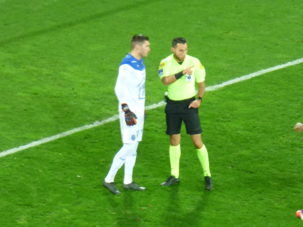 Ein Schiedsrichter in einem gelben T-Shirt und schwarzen Shorts spricht mit einem Fußballspieler in einem weißen und blauen Trikot auf dem Feld, beide tragen Turnschuhe.