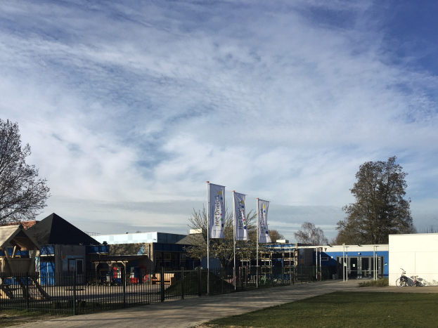 Großes Gebäude mit einem großen Grasbereich davor, umgeben von einem Metallzaun, mit einem Weg, Bäumen, Pfosten, Bannern, einem geparkten Fahrrad, nahegelegenen Häusern und einem bewölkten Himmel, identifiziert als der neue Spielplatz der Schule.
