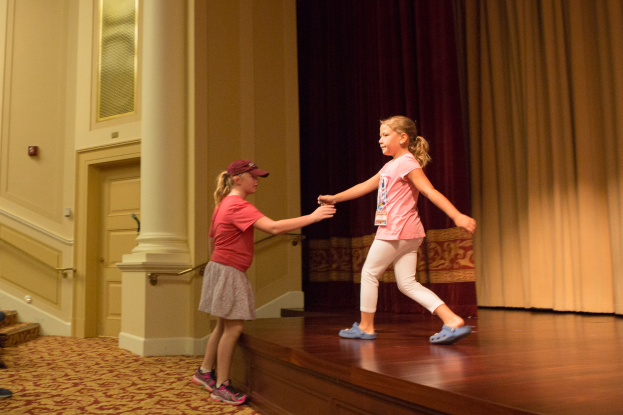Zwei junge Mädchen tanzen auf einer Bühne vor einem Vorhang, mit einer Person, die links von ihnen auf einem Stuhl sitzt, einer Säule und einer Wand im Hintergrund, beide Mädchen tragen Schuhe.