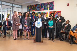 Eine Gruppe von Menschen, die vor einem Podium mit Mikrofon und einer Tafel mit Text darauf stehen und sitzen. Im Hintergrund sind Flaggen, eine Tafel, ein Feuerl├Âscher und einige Deckenleuchten zu sehen. Der Text auf der Tafel lautet "Sancho City Councilman Joins City Council".