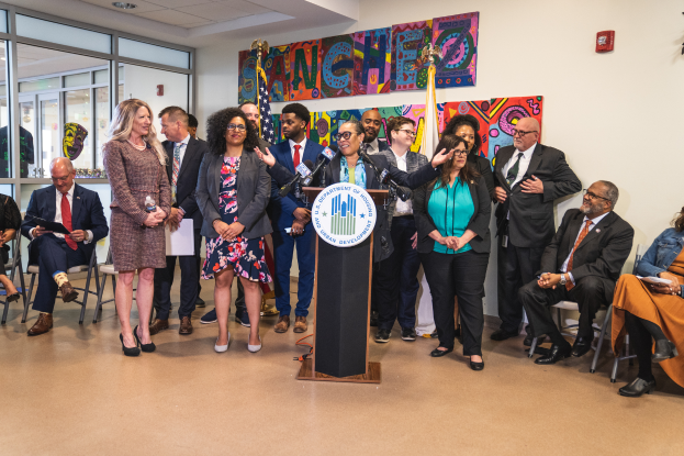 Eine Gruppe von Menschen, die vor einem Podium mit Mikrofon und einer Tafel mit Text darauf stehen und sitzen. Im Hintergrund sind Flaggen, eine Tafel, ein Feuerl├Âscher und einige Deckenleuchten zu sehen. Der Text auf der Tafel lautet "Sancho City Councilman Joins City Council".