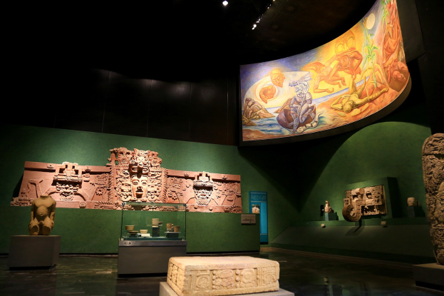 Ein Museumsraum mit verschiedenen Artefakten, darunter Skulpturen, Objekte auf Tischen, eine Texttafel, eine Wandmalerei und Deckenbeleuchtung im Anthropologischen Museum von Mexiko-Stadt.