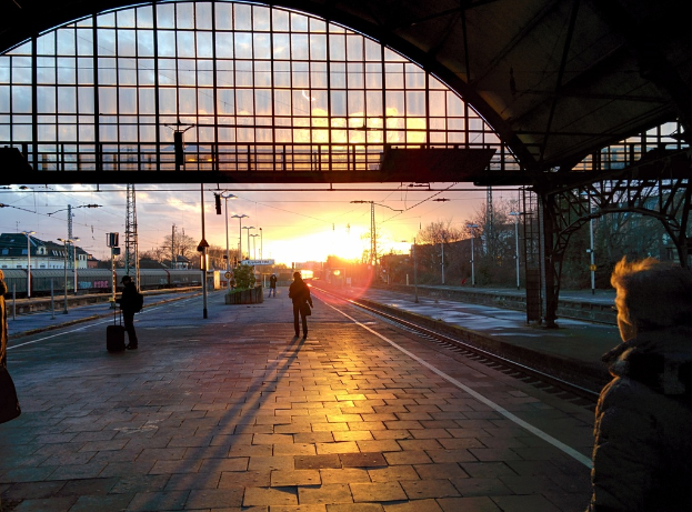Ein Bahnsteig mit wenigen Menschen, ein Zug auf der linken Seite, zahlreiche Pfosten, Sonnenstrahlen, Bäume auf der rechten Seite und ein Dach darüber.