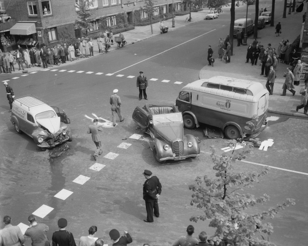 Schwarz-weißes Bild einer Autounfall-Szene an einer Straßenkreuzung mit einem Van, anderen Fahrzeugen und einer Gruppe von Menschen, die herumstehen, mit Gebäuden, Bäumen, Polen und Texttafeln im Hintergrund.