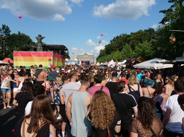 Eine große Menge geht eine Straße mit Zelten, Bäumen, Pfählen, Lichtern und einer Statue entlang während des Christopher Street Day in Berlin, mit Gebäuden, Wolken und Ballons im Hintergrund.