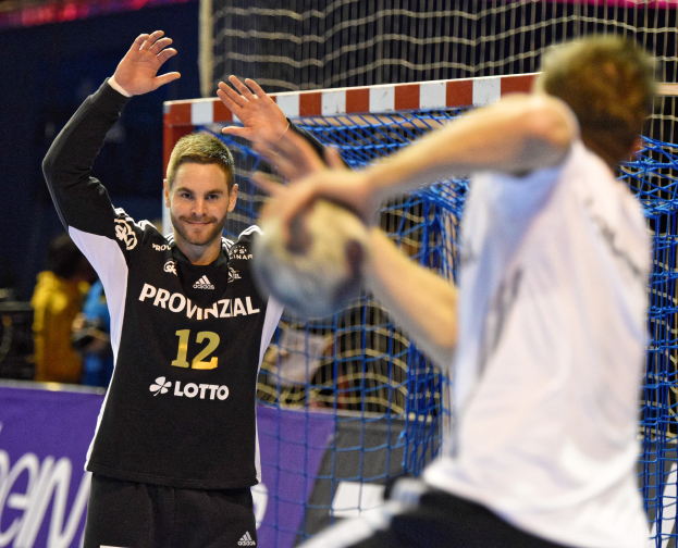 Zwei Männer spielen Handball vor einem Tor mit Netz und einem Banner mit Text im Hintergrund und einigen Zuschauern in einer Stadion.