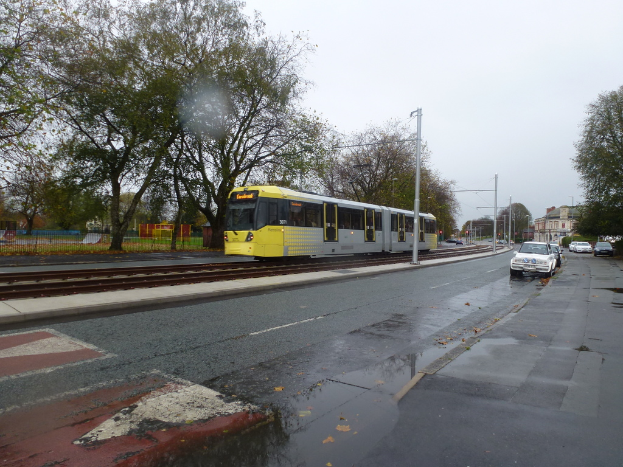 Eine belebte Straßenszene mit Fahrzeugen, Gebäuden, Pfählen, einer Bahnstrecke mit einem Zug, Bäumen und einem Zaun unter einem klaren Himmel.