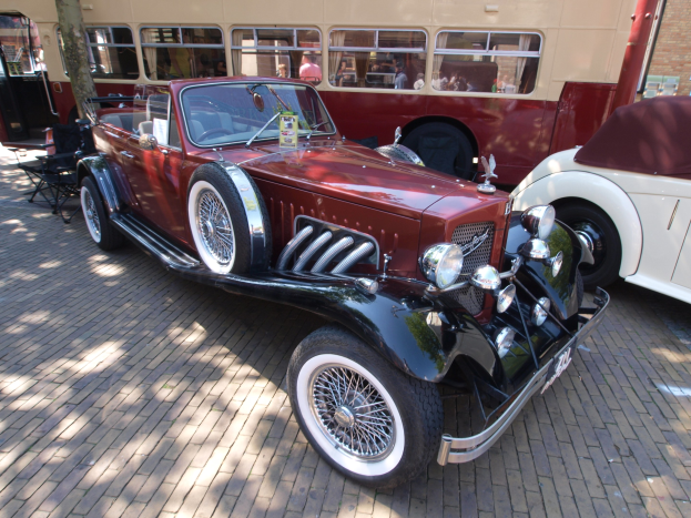 Vintage-Auto neben einem Doppeldeckerbus auf Kopfsteinpflasterstraße geparkt, mit Stühlen und Baum im Vordergrund, einem Gebäude mit Fenstern im Hintergrund und Menschen im Bus zu sehen.