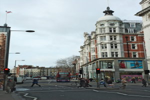 Ein roter Doppeldeckerbus fährt eine belebte Stadtstraße entlang, die von hohen Gebäuden, Laternen, Ampeln und Menschen auf dem Gehweg sowie Bäumen im Hintergrund und einem klaren blauen Himmel gesäumt ist.