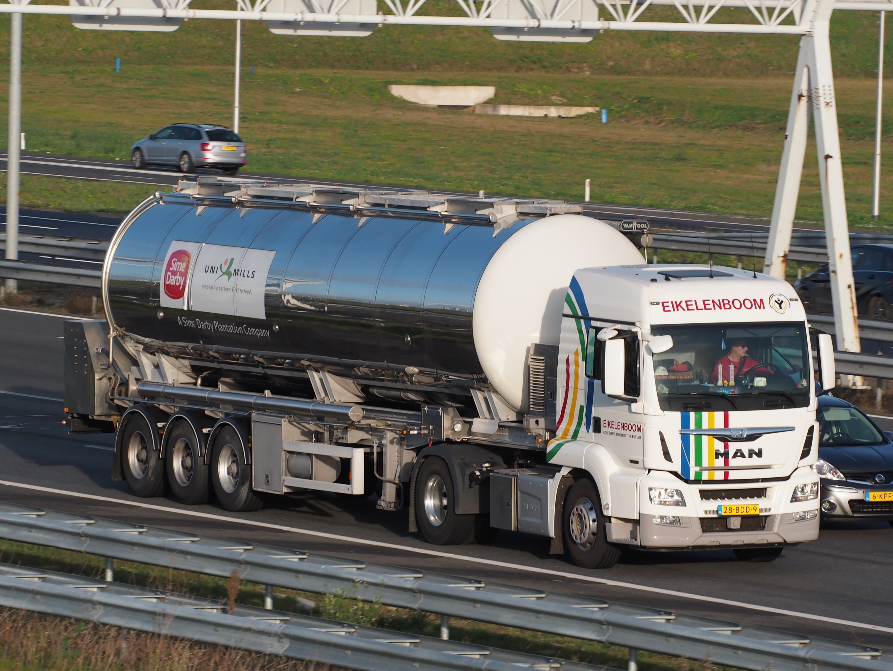 Großer Tanklastwagen auf einer Autobahn neben einem Auto mit sichtbaren Insassen im Truck und Gras, Stangen und anderen Gegenständen im Hintergrund.