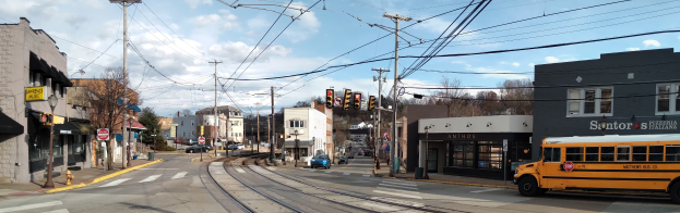 Ein gelber Schulbus fährt auf einer Straße mit hohen Gebäuden, Strommasten, Ampeln, Schildern, Bäumen und einem bewölkten Himmel im Hintergrund.