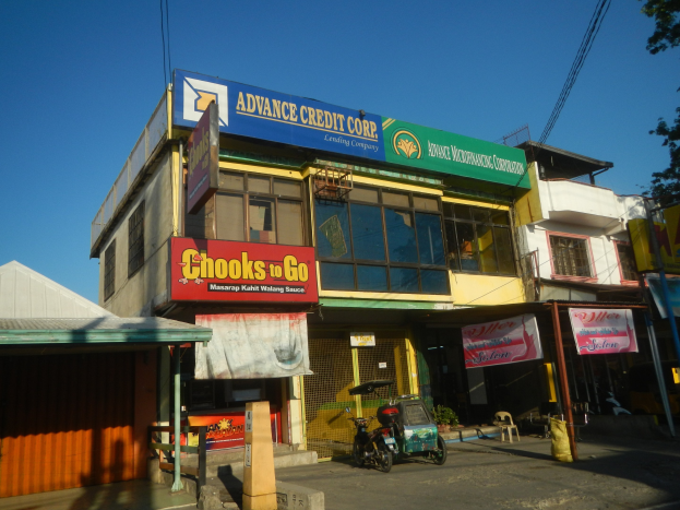 Ein Geschäftsgebäude mit einer Tafel, auf der "Advance Credit Corp" steht, an einer vielbefahrenen Straße mit Fahrzeugen, Werbetafeln, Strommasten und Bäumen unter einem klaren Himmel.