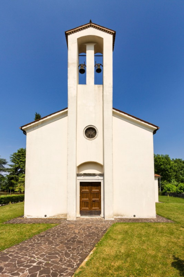 Eine kleine weiße Kirche mit einem Glockenturm, umgeben von einem Weg, Gras, Pflanzen und Bäumen, unter einem bewölkten Himmel, mit laut klingelnder Glocke.
