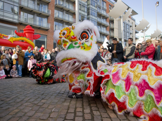 Ein farbenfrohes chinesisches Neujahrsfest in Amsterdam mit einer Löwen-Tanz-Performance vor einer Zuschauermenge, einige halten Kameras, vor einem Hintergrund aus Gebäuden, Laternenmasten und einem klaren blauen Himmel.
