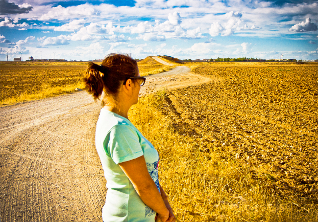 Eine Frau steht auf einem schlammigen Weg, der von landwirtschaftlichen Flächen und Bäumen gesäumt ist.